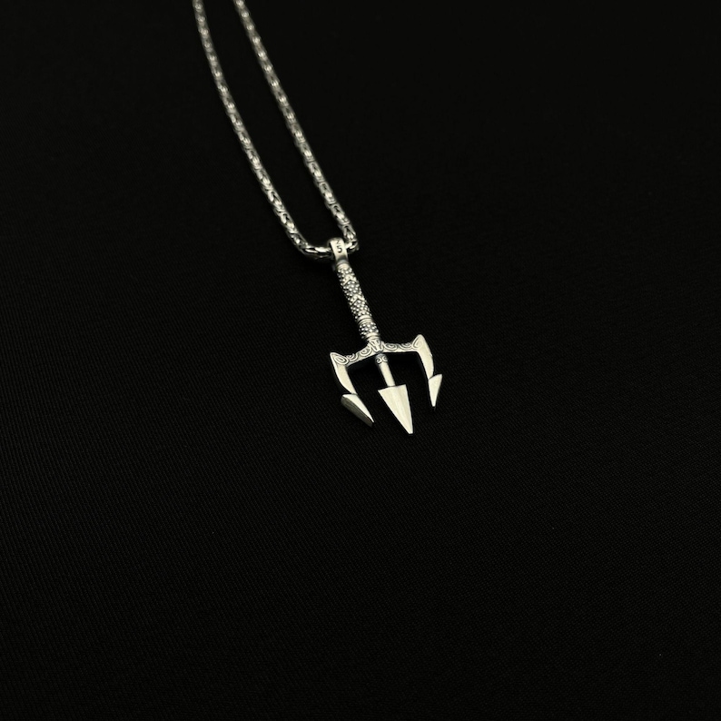 May include: A silver-colored pendant necklace featuring a trident design. The trident has three sharp prongs and is suspended from a delicate chain. The necklace is displayed against a black background, highlighting the intricate details of the jewelry.