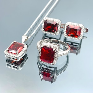 May include: A silver necklace, ring, and earrings set with red cubic zirconia stones. The jewellery features a square-shaped stone with a silver halo setting.