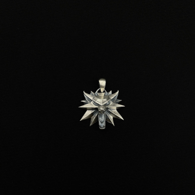 May include: A silver pendant featuring a stylized wolf's head with sharp, angular features. The pendant has a loop at the top for a chain and is set against a black background. The wolf's head has an open mouth and pointed ears.