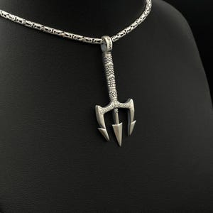 May include: A silver trident pendant hangs from a silver chain. The pendant has a detailed design with three prongs and a decorative handle. The chain has a textured, woven pattern. The pendant is likely made of sterling silver.