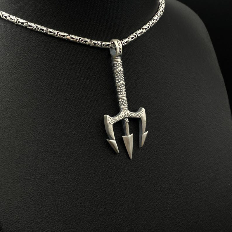 May include: A silver trident pendant hangs from a silver chain. The pendant has a detailed design with three prongs and a decorative handle. The chain has a textured, woven pattern. The pendant is likely made of sterling silver.