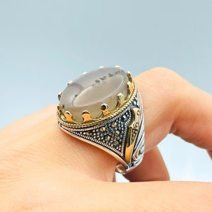 Pode incluir: Um anel de prata com uma pedra de &aacute;gata cinza engastada em uma coroa dourada. O anel &eacute; decorado com um design detalhado e pedras pretas de marcasita.