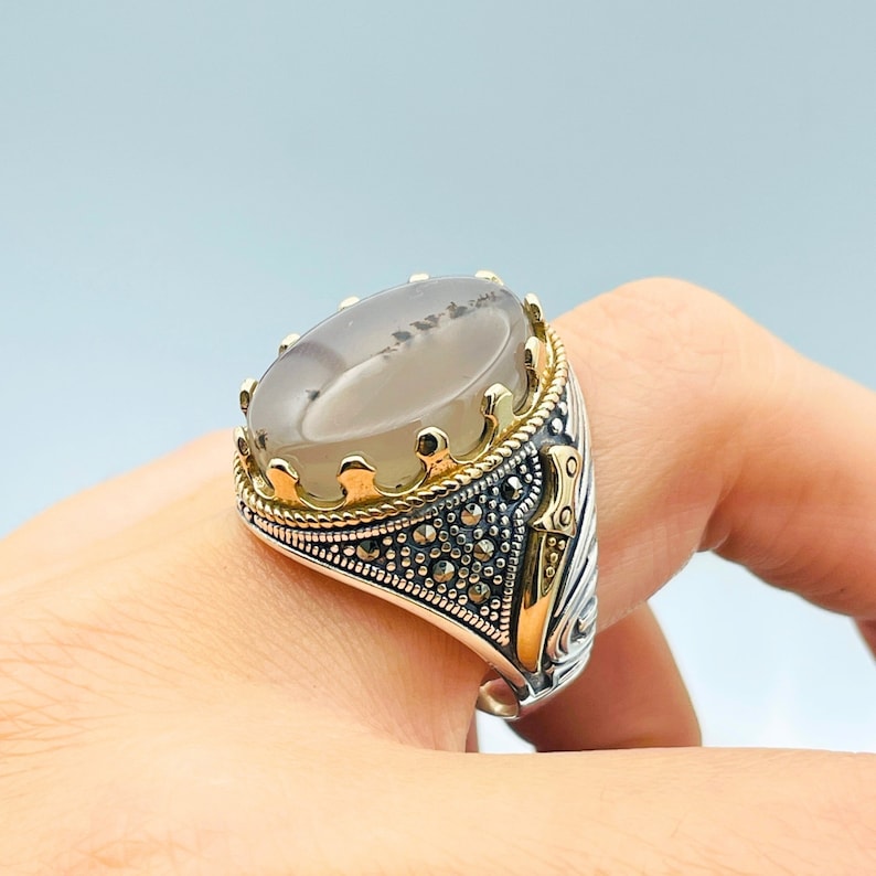 Pode incluir: Um anel de prata com uma pedra de &aacute;gata cinza engastada em uma coroa dourada. O anel &eacute; decorado com um design detalhado e pedras pretas de marcasita.