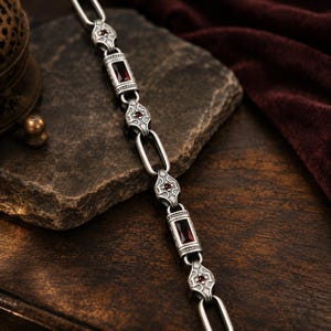 May include: A silver-toned chain bracelet featuring alternating rectangular links with deep red stones and ornate, shield-shaped links accented with tiny red gems. The metal has a darkened, oxidized finish, creating a high-contrast look.