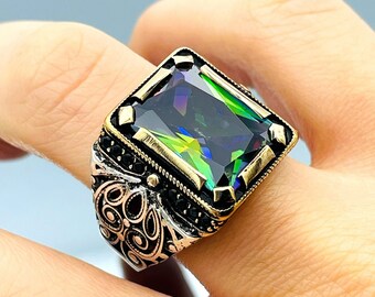 May include: A silver ring with a large, emerald-cut gemstone. The gemstone has a dark green base with iridescent rainbow hues. The ring has a detailed, ornate design with black accents.