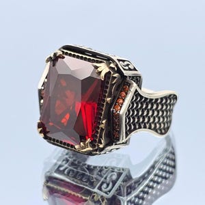 May include: A silver ring with a large, rectangular, faceted, deep red gemstone. The ring has intricate gold-colored filigree and small orange-red accent stones. The band features a textured, scale-like pattern.