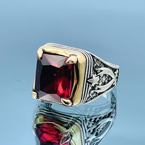 May include: A silver ring with a large, rectangular, deep red gemstone. The ring has gold accents around the stone and intricate detailing on the band, including a star and crescent moon design. The ring is displayed on a reflective surface.