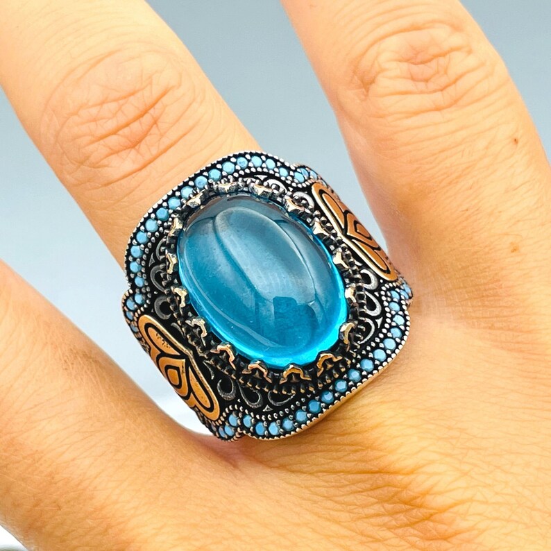 May include: A silver ring with a large oval blue stone set in a detailed filigree design. The ring is accented with small blue stones around the edge.