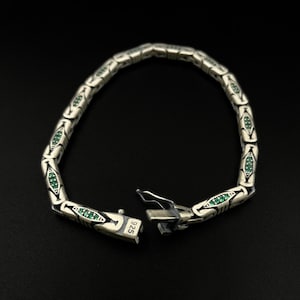May include: Silver bracelet with a repeating pattern of stylized shapes, each containing small green gemstones. The bracelet is unclasped, revealing the clasp mechanism. The clasp is stamped with "925". The bracelet is displayed against a black background.
