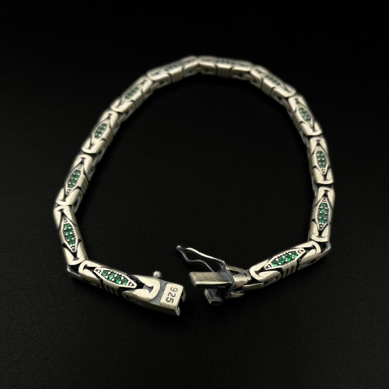 May include: Silver bracelet with a repeating pattern of stylized shapes, each containing small green gemstones. The bracelet is unclasped, revealing the clasp mechanism. The clasp is stamped with "925". The bracelet is displayed against a black background.