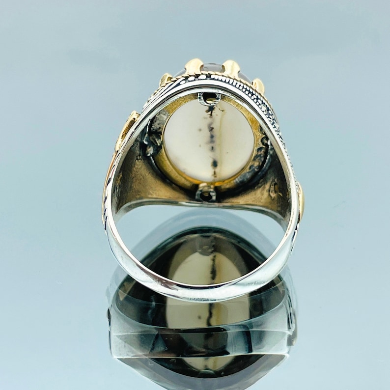 Pode incluir: Um anel de prata com uma grande pedra ovalada engastada em uma moldura de ouro. O anel &eacute; decorado com detalhes intrincados e tem uma banda de prata.