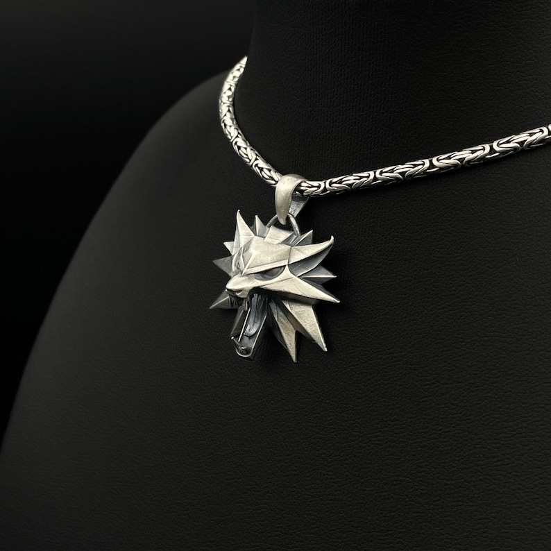 May include: A silver pendant necklace featuring a stylized wolf head emblem. The pendant has a geometric design with sharp angles and a detailed face. The necklace chain is a textured, woven style. The jewelry is displayed against a black background.