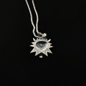 May include: A silver pendant necklace featuring a sunburst design with a central geometric shape. The pendant is attached to a delicate chain. The background is solid black.