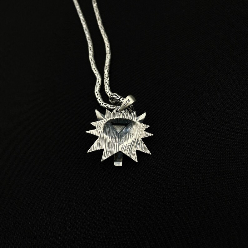 May include: A silver pendant necklace featuring a sunburst design with a central geometric shape. The pendant is attached to a delicate chain. The background is solid black.
