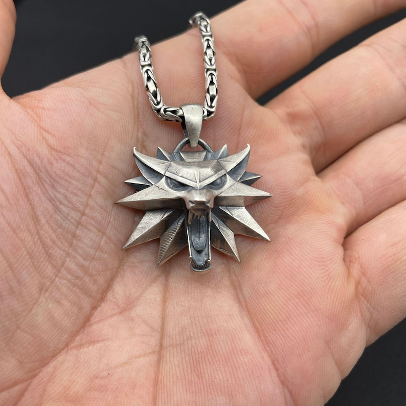 May include: A silver-colored pendant necklace featuring a wolf's head emblem. The pendant has a detailed, angular design with the wolf's mouth open. The necklace chain is a textured, linked design.