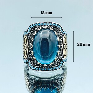 May include: A silver ring with a large blue gemstone surrounded by smaller blue stones. The ring is 15 mm wide and 20 mm tall.