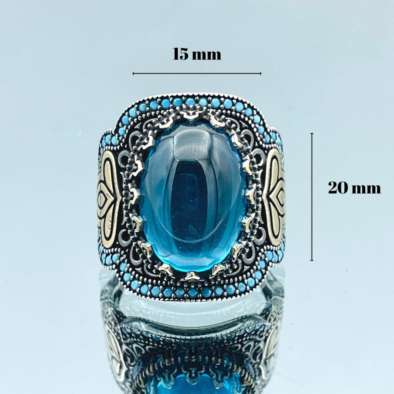 May include: A silver ring with a large blue gemstone surrounded by smaller blue stones. The ring is 15 mm wide and 20 mm tall.