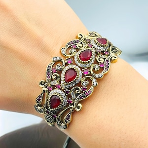 Puede incluir: Un brazalete de tono dorado con un diseño filigranado detallado. El brazalete está adornado con piedras preciosas rojas y transparentes.