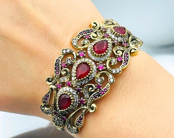 Könnte beinhalten: Ein goldfarbenes Armband mit einem detaillierten Filigranmuster. Das Armband ist mit roten und klaren Edelsteinen verziert.