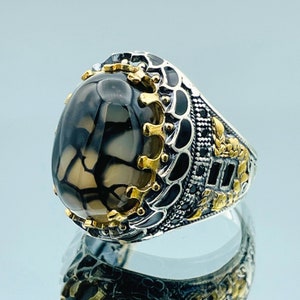 May include: A silver ring with a black and brown agate stone set in a gold crown design. The ring band is detailed with intricate silver and gold patterns.