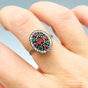 May include: A silver ring with an oval-shaped design featuring a large red gemstone surrounded by smaller green and pink gemstones.
