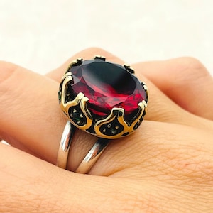 Può includere: Un anello d'argento con una grande pietra preziosa rossa incastonata in una montatura dorata e decorata. L'anello è su un dito.