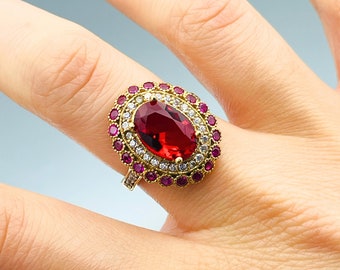 Könnte beinhalten: Ein goldener Ring mit einem großen ovalen roten Edelstein, umgeben von kleineren klaren und roten Edelsteinen.