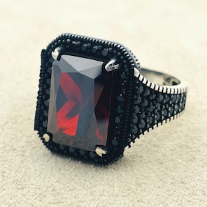 May include: A silver ring with a large, dark red gemstone set in a black bezel. The ring is decorated with small black stones around the band.
