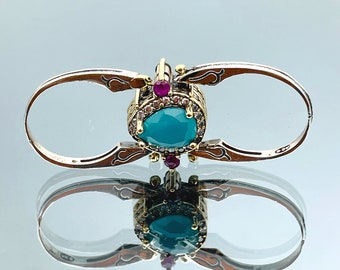 May include: A silver ring with a turquoise gemstone surrounded by smaller clear and pink gemstones. The ring is designed to be worn on two fingers.