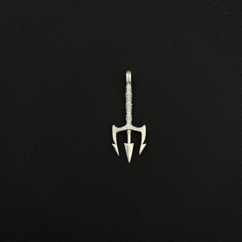 May include: A silver trident pendant with a detailed design. The pendant has a three-pronged head and a textured handle. The trident is displayed against a black background.