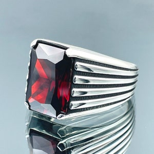 May include: A silver ring with a large, rectangular, deep red gemstone. The ring has a ribbed design on the sides, and the gemstone is faceted, catching the light. The ring is displayed on a reflective surface.