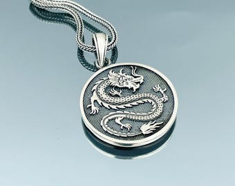 Peut inclure: Un collier avec un pendentif en argent représentant un disque rond orné d'un dragon argenté détaillé. Le dragon est enroulé et présente une apparence texturée et écailleuse.