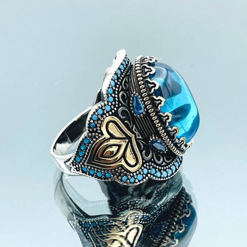May include: A silver ring with a large blue gemstone set in a gold crown. The ring is decorated with intricate filigree and smaller blue gemstones.