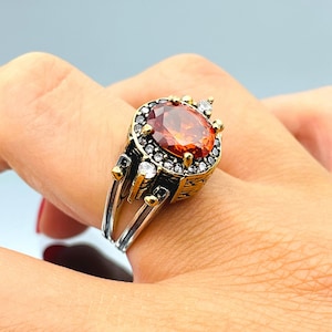May include: A silver and gold ring featuring a large orange gemstone surrounded by smaller clear gemstones. The ring has an elaborate design with intricate carvings.