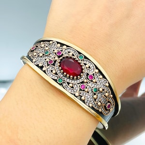 May include: A silver bracelet with a gold-toned design featuring a large red gemstone surrounded by smaller green and pink gemstones.