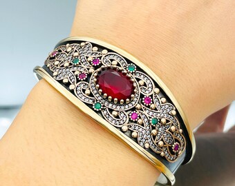 Könnte beinhalten: Ein silbernes Armband mit einem goldfarbenen Design, das einen großen roten Edelstein zeigt, der von kleineren grünen und rosa Edelsteinen umgeben ist.