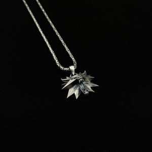 May include: A silver-colored pendant necklace featuring a stylized wolf head emblem. The pendant has a detailed, angular design with sharp points and a textured surface. The chain is a linked design, and the background is black.