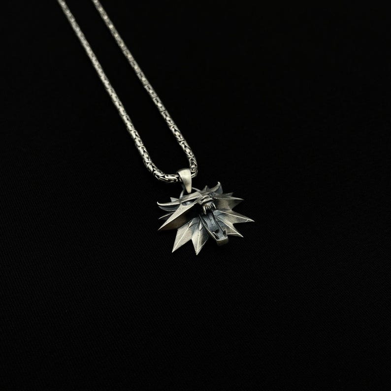 May include: A silver-colored pendant necklace featuring a stylized wolf head emblem. The pendant has a detailed, angular design with sharp points and a textured surface. The chain is a linked design, and the background is black.