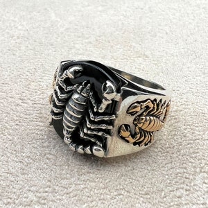 May include: A silver ring with a black onyx stone and scorpion design. The ring features a detailed scorpion on the front and side, with gold-colored accents. The ring has a wide band and a polished finish.