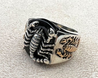 Puede incluir: Un anillo de plata con una piedra de ónix negro y un diseño de escorpión. El anillo presenta un escorpión detallado en la parte delantera y lateral, con detalles dorados. El anillo tiene una banda ancha y un acabado pulido.