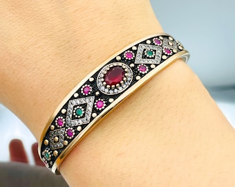 Pode incluir: Um bracelete de prata com acabamento oxidado preto. O bracelete é decorado com um padrão geométrico de diamantes e pedras preciosas vermelhas e verdes.