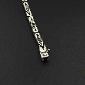 May include: A silver bracelet with a repeating pattern of stylized shapes, each containing small green gemstones. The clasp is marked with "925". The bracelet is displayed against a dark background.