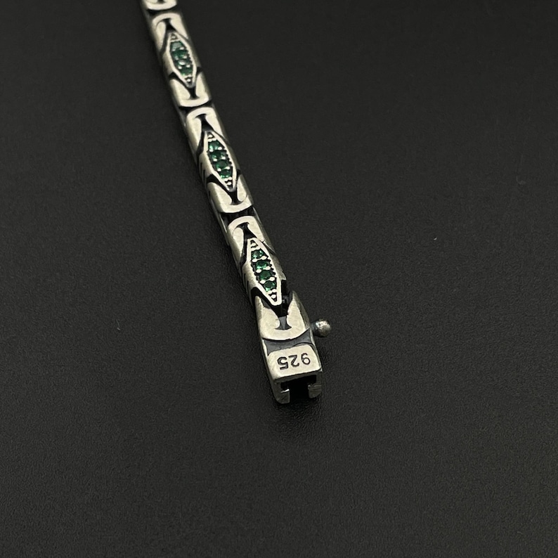May include: A silver bracelet with a repeating pattern of stylized shapes, each containing small green gemstones. The clasp is marked with "925". The bracelet is displayed against a dark background.