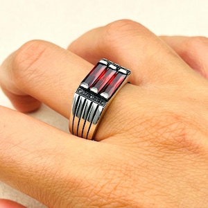 May include: A silver ring with a rectangular design featuring three red gemstones and black accents.