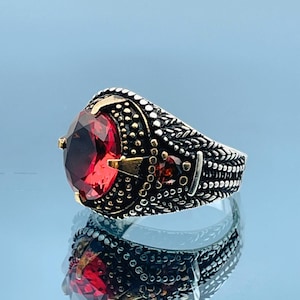 May include: A silver ring with a large, oval, deep red gemstone as the centerpiece. The ring has intricate detailing with gold accents and a textured band, creating a vintage or antique aesthetic. The gemstone is the focal point.