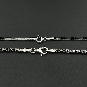 May include: Close-up of two silver necklaces with different chain styles. The top necklace has a fine, textured chain with a small clasp. The bottom necklace features a more ornate, patterned chain with a larger clasp. Both are on a dark background.