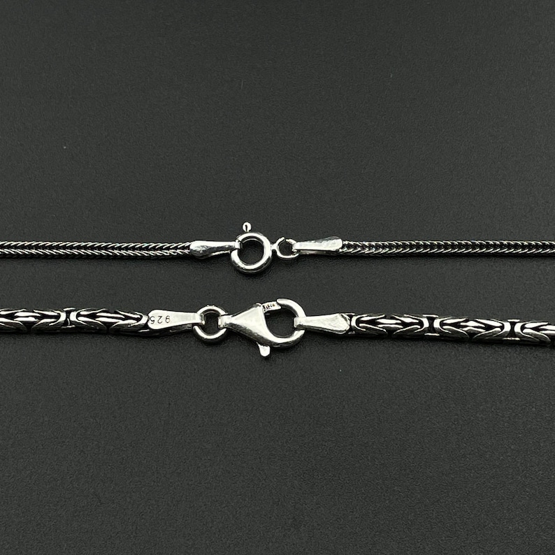 May include: Close-up of two silver necklaces with different chain styles. The top necklace has a fine, textured chain with a small clasp. The bottom necklace features a more ornate, patterned chain with a larger clasp. Both are on a dark background.