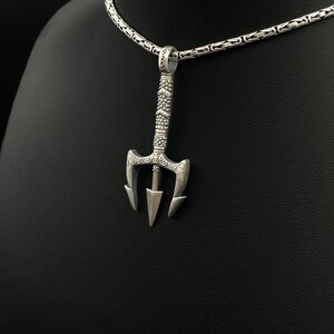 May include: A silver trident pendant with intricate detailing hangs from a silver chain. The pendant features a three-pronged design with decorative patterns on the handle. The chain is a textured, linked style.