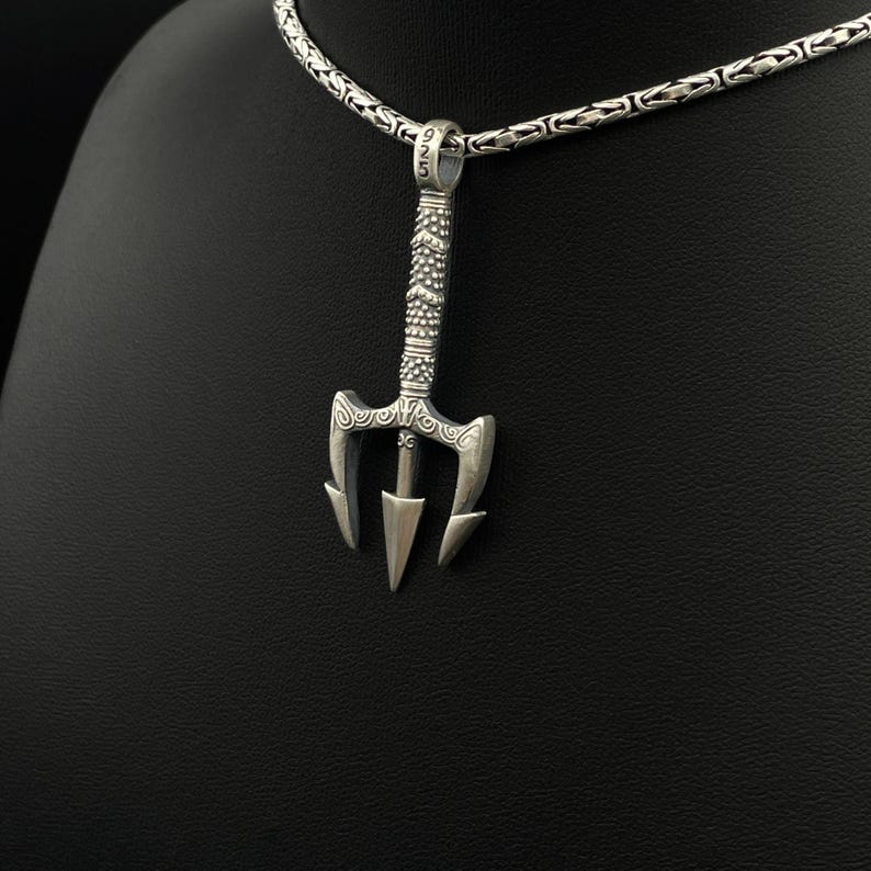 May include: A silver trident pendant with intricate detailing hangs from a silver chain. The pendant features a three-pronged design with decorative patterns on the handle. The chain is a textured, linked style.