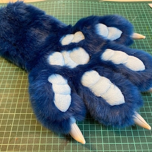 May include: Blue furry paw with light blue pads and white claws. The paw is lying on a green cutting mat.